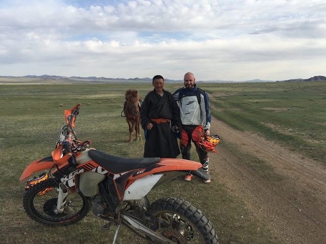 Two people standing beside motorcycles on a vast plain with a horse in the background.