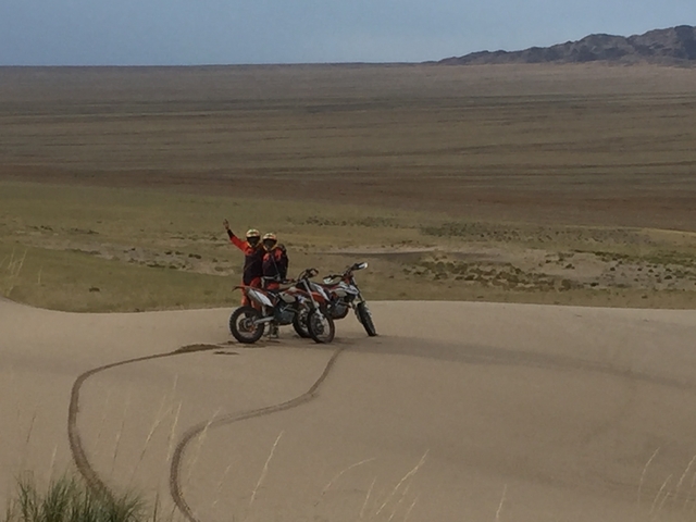 Two people on motorbikes on sand dunes with one raising an arm in a gesture.