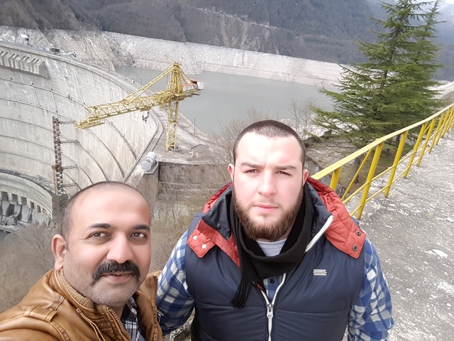 Two people taking a selfie with a large dam in the background.