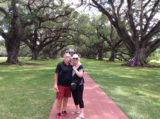       Couple posing under oak trees leading to a house.
  