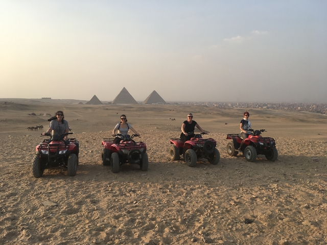 Group of people riding ATVs in a desert with pyramids in the background.