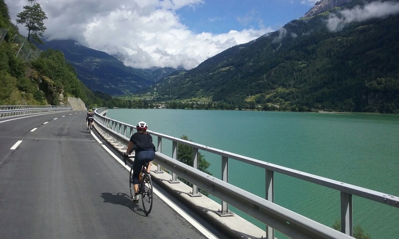 Cyclists riding along a scenic lakeside road with mountain views.