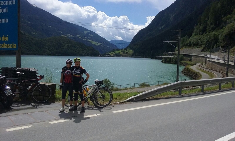 Two cyclists posing by a scenic lakeside road with bicycles.