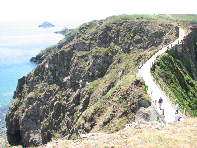 A rugged coastal path with people walking, cliffs, and the ocean.