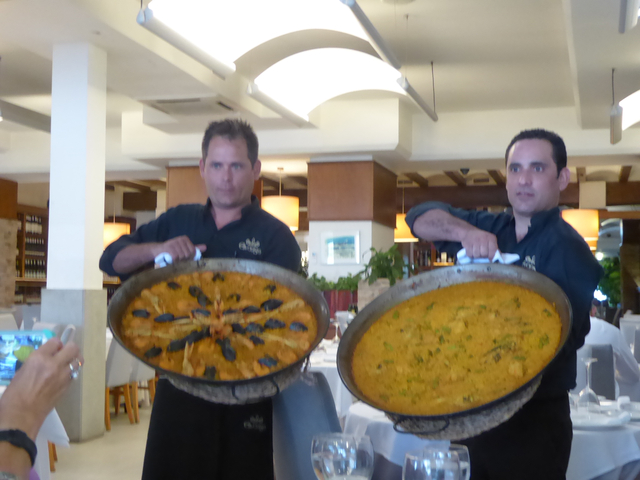 Two waiters holding large pans of paella indoors.