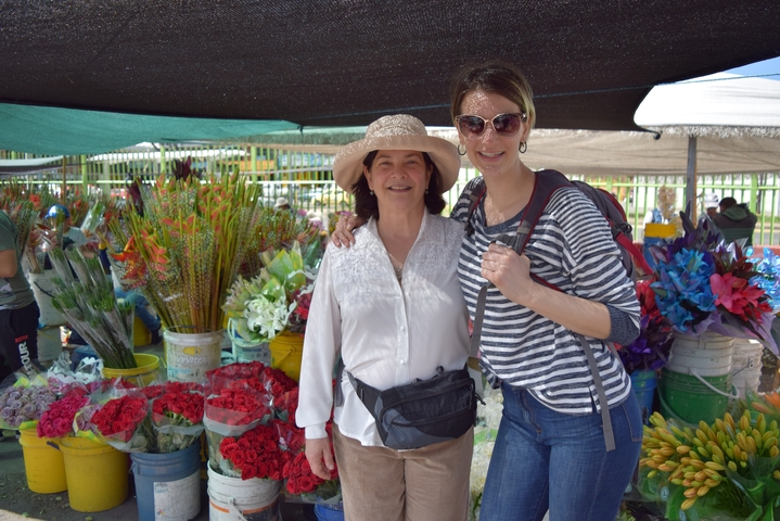 Two women smiling at a market with an abundance of colorful flowers.