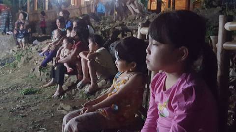       Children and adults seated outdoors watching an event at night.
  