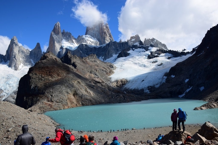       Two hikers near a glacial lake with mountains.
  