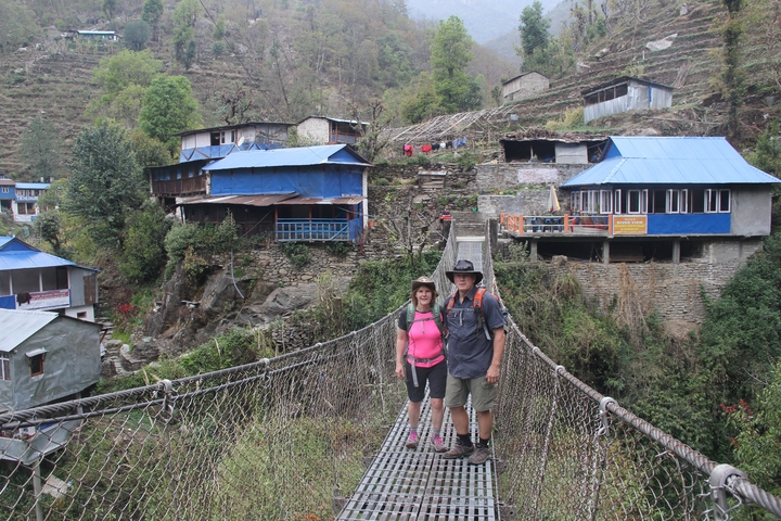 Two hikers on a suspension bridge in a hilly village landscape.