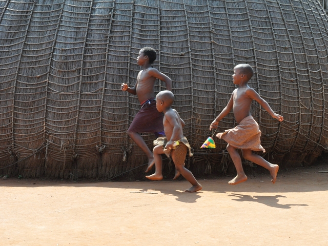 Three children joyfully running along a traditional thatched wall.