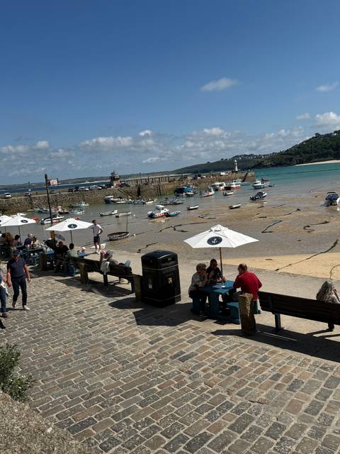 Harbor with boats and people sitting on a bench with an umbrella and stone pavement.