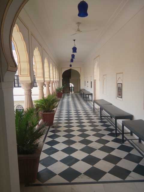 An interior view of a corridor with checkerboard flooring and potted plants.