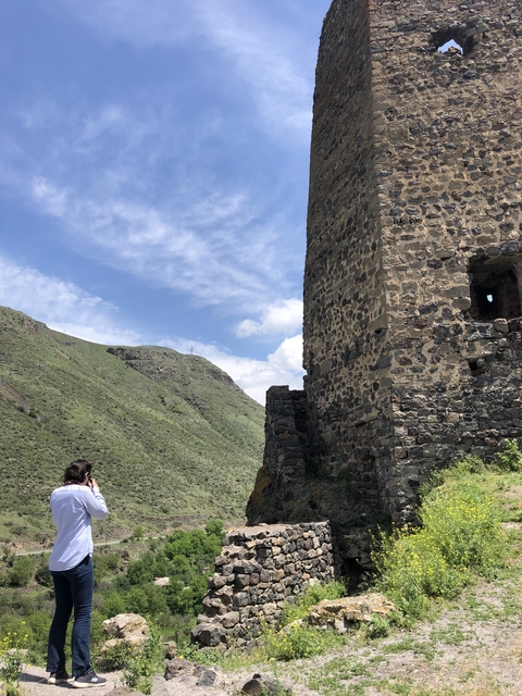       A person photographing an ancient stone building with mountains in the background.
  