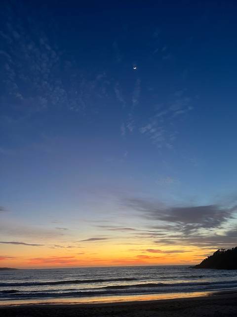Colorful sunset over the ocean with a crescent moon in the sky.