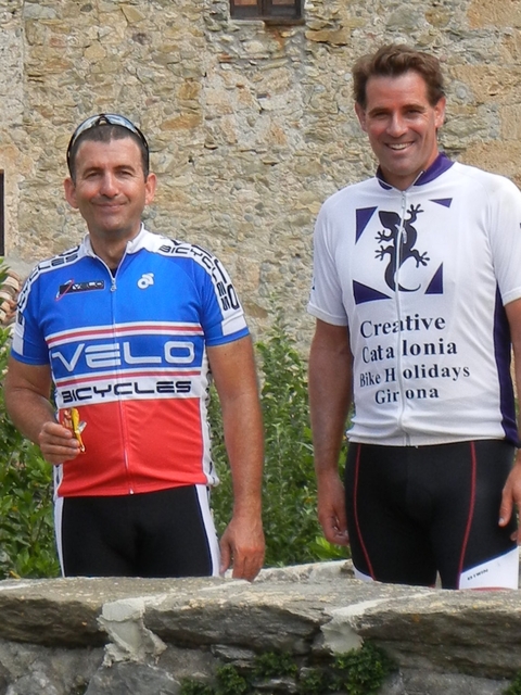 Two cyclists in branded jerseys standing with a wall in the background.