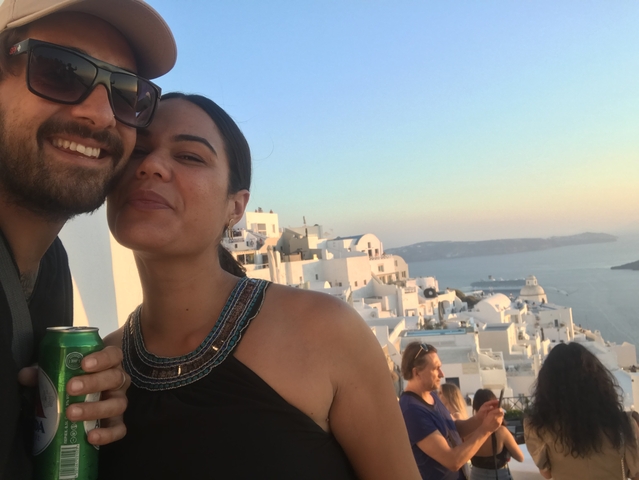       Couple at a scenic viewpoint with the sea and white buildings in the background.
  
