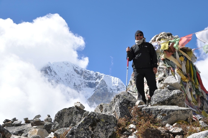 Person posing on rocky terrain with snowy mountains and prayer flags in the background.