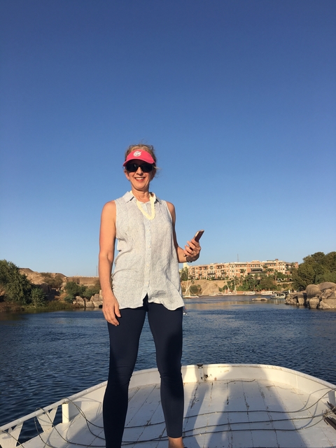       Woman standing by a river with buildings in the background, dressed casually.
  
