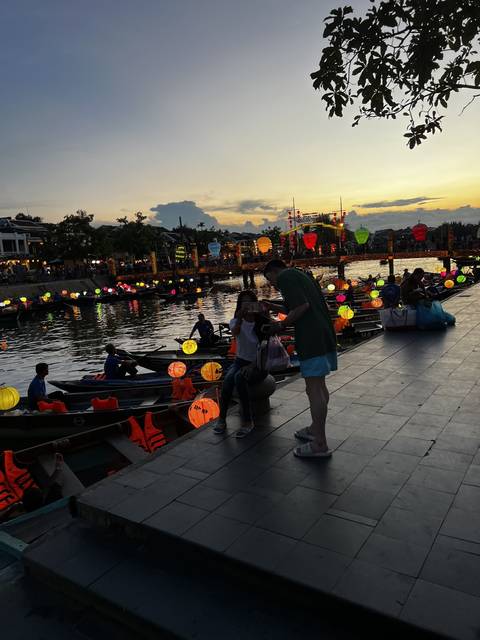 People by a river with boats adorned with colorful lanterns in the evening.