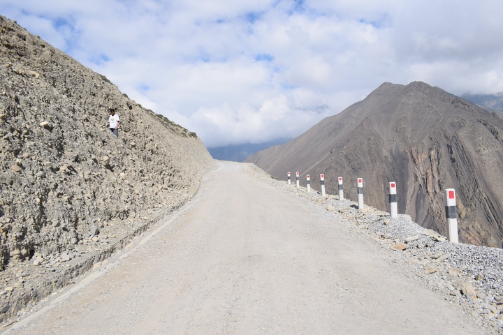 A mountain road with a person walking on the left side, surrounded by rugged hills and a cloudy sky.