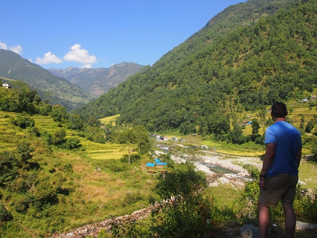       A person on a hilltop overlooking a lush green valley with scattered houses and distant mountains.
  