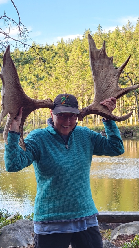       Person posing playfully with moose antlers near a lakeside.
  