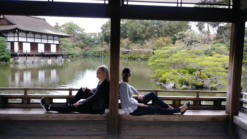       Two women seated back-to-back in a wooden pavilion by a pond.
  