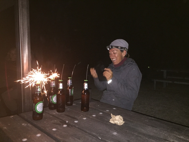      A person with sparklers and beer bottles on a picnic table at night.
  