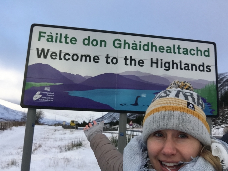 Person posing by a sign reading 'Welcome to the Highlands'.