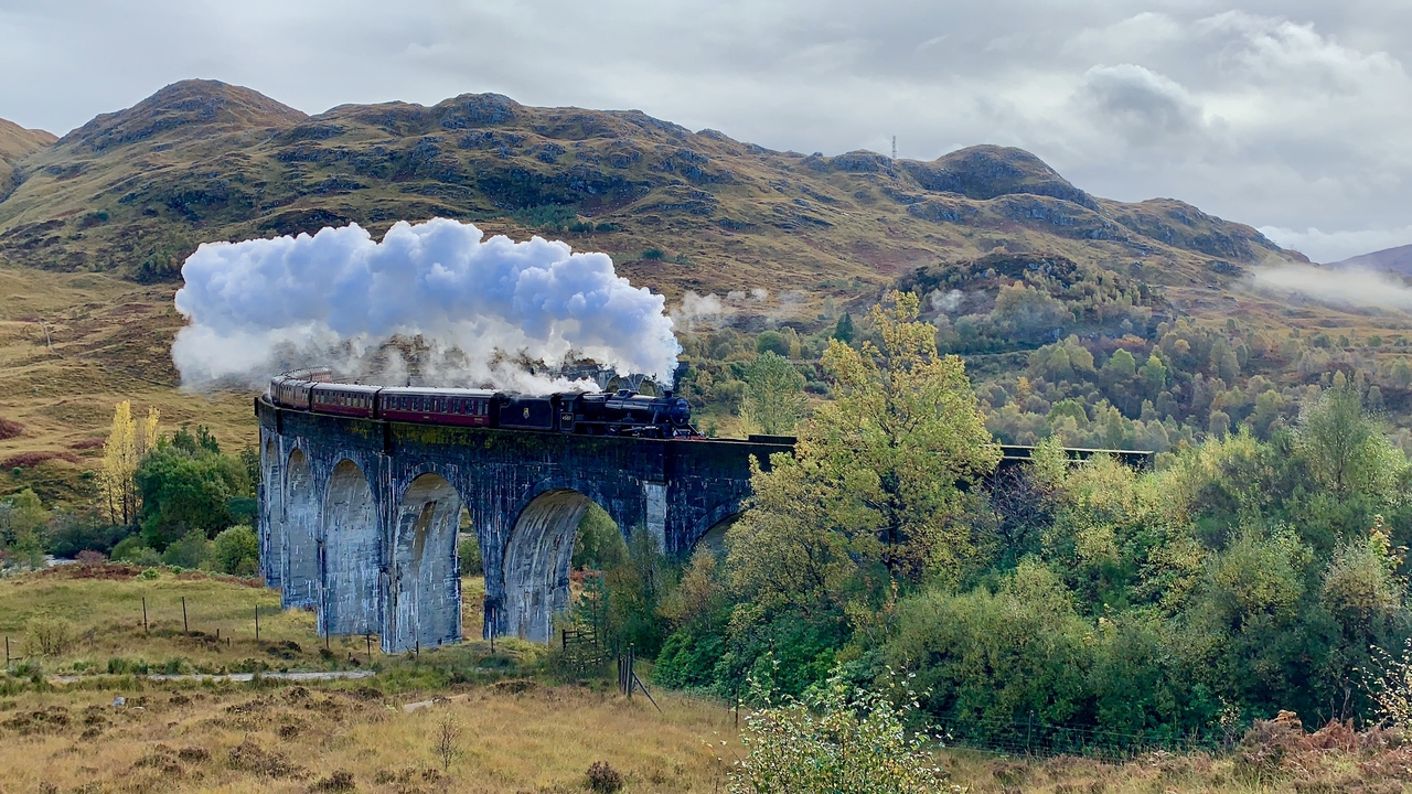 Steam train crossing a viaduct in a scenic landscape with hills and forests.