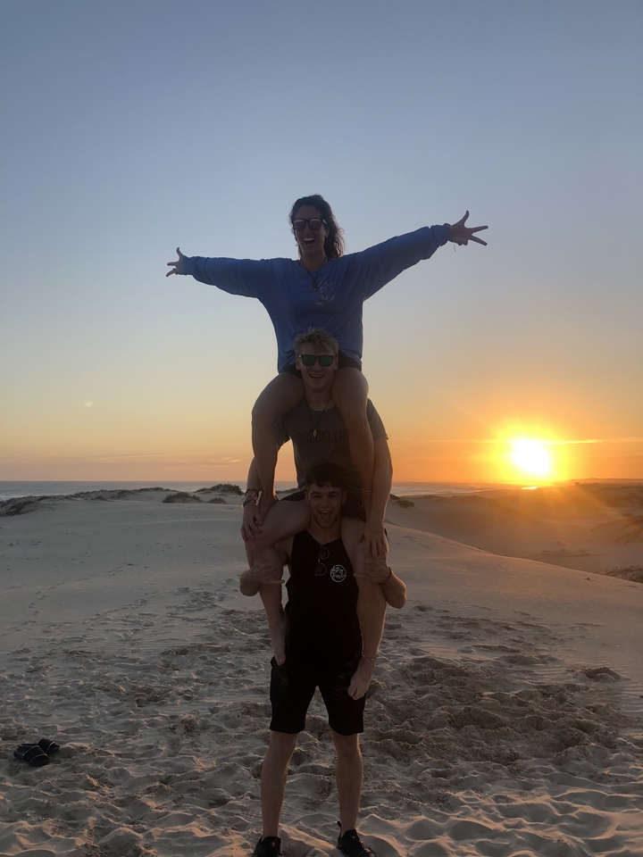 Three people stacked in a playful pose on a sandy beach at sunset.