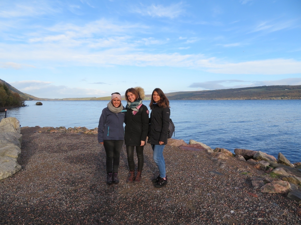 Three women standing by a lake with hills in the background.