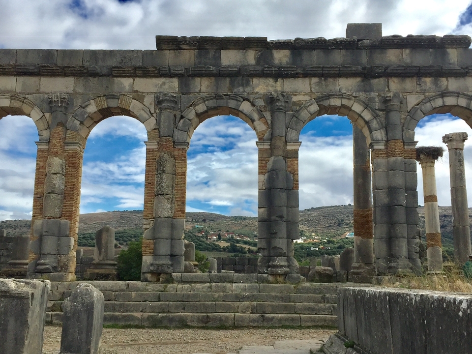 Ancient arcades with a mountainous backdrop under a cloudy sky.