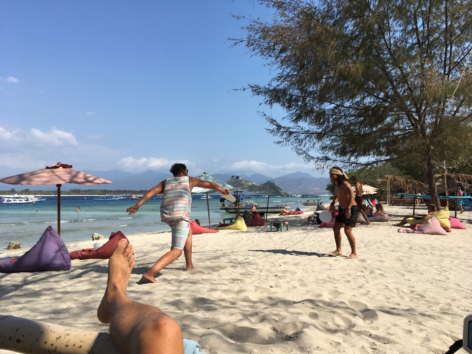 People playing on a beach with bean bags and umbrellas, mountains in the background.