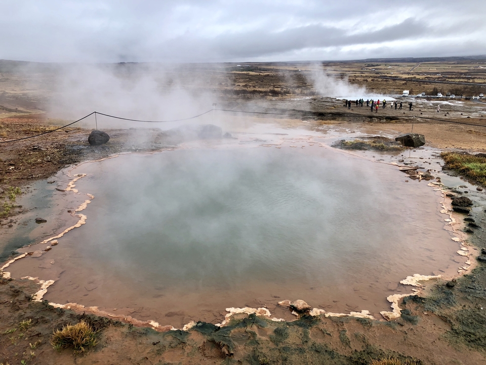 Hot spring with steam rising, surrounded by rocky terrain.
