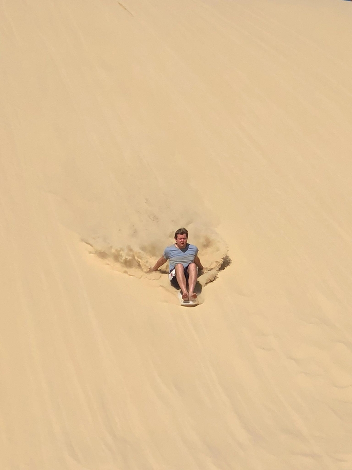 Man sliding down a large sand dune with sand spraying around.