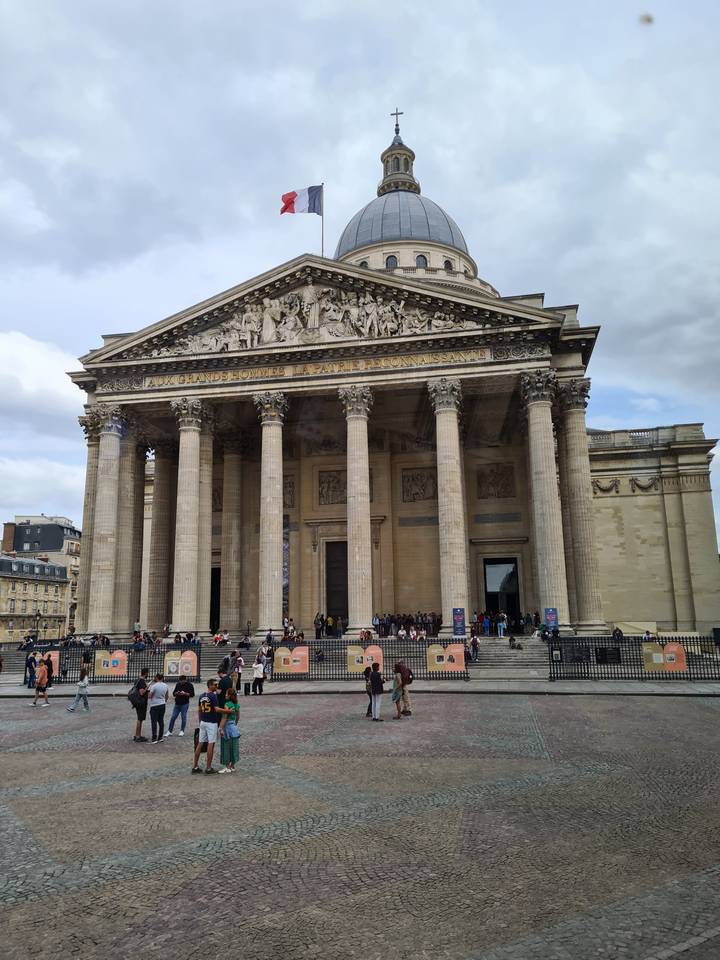 A group of people in front of a grand building with columns and a dome, French flag visible.