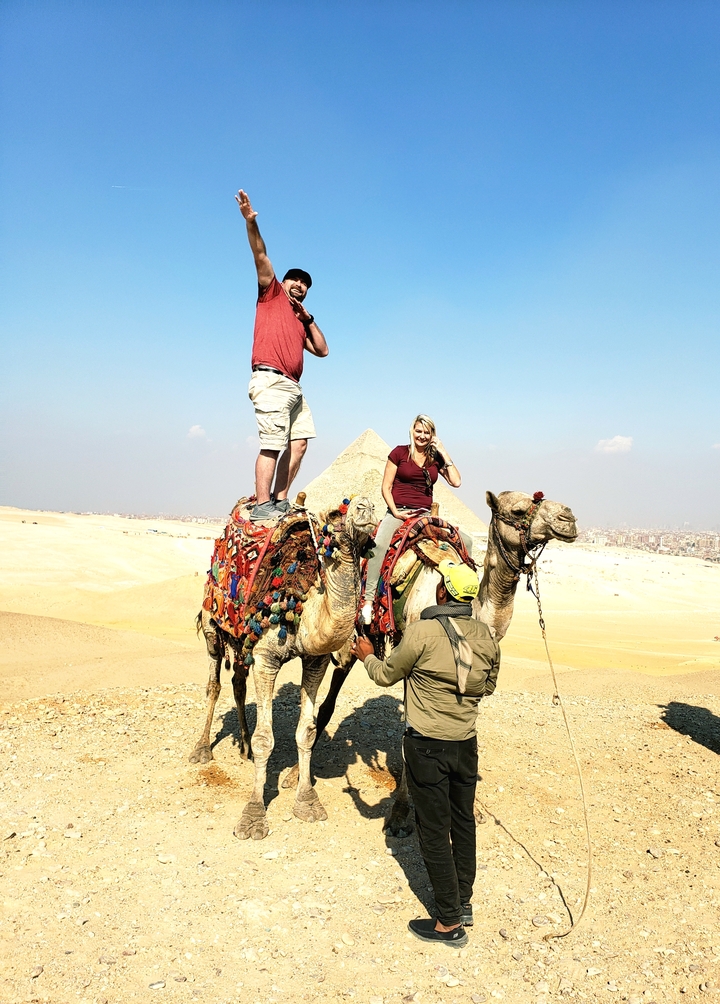 Two people on camels with pyramids in the background.