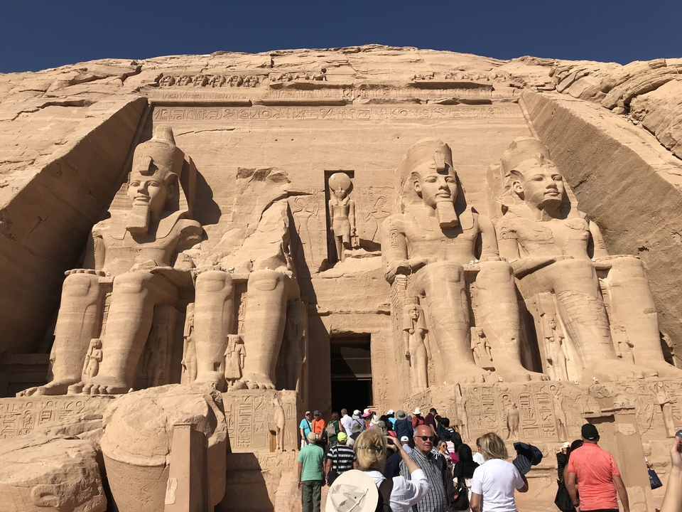 Large statues carved into a sandstone cliff with tourists.