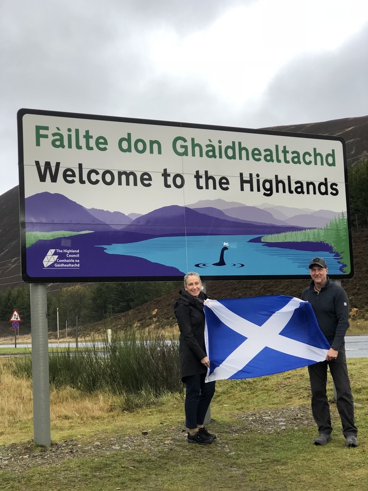Two people posing with a Scottish flag next to a welcome sign.