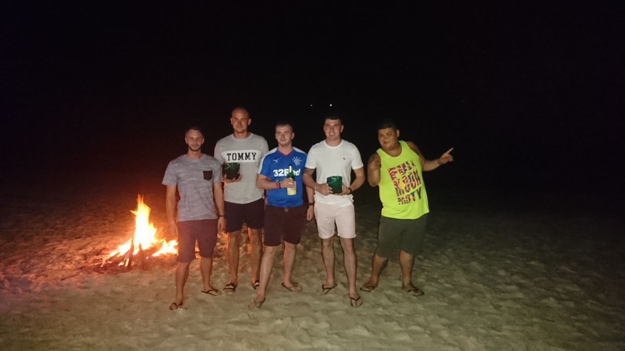 Group of men standing on the beach near a campfire.