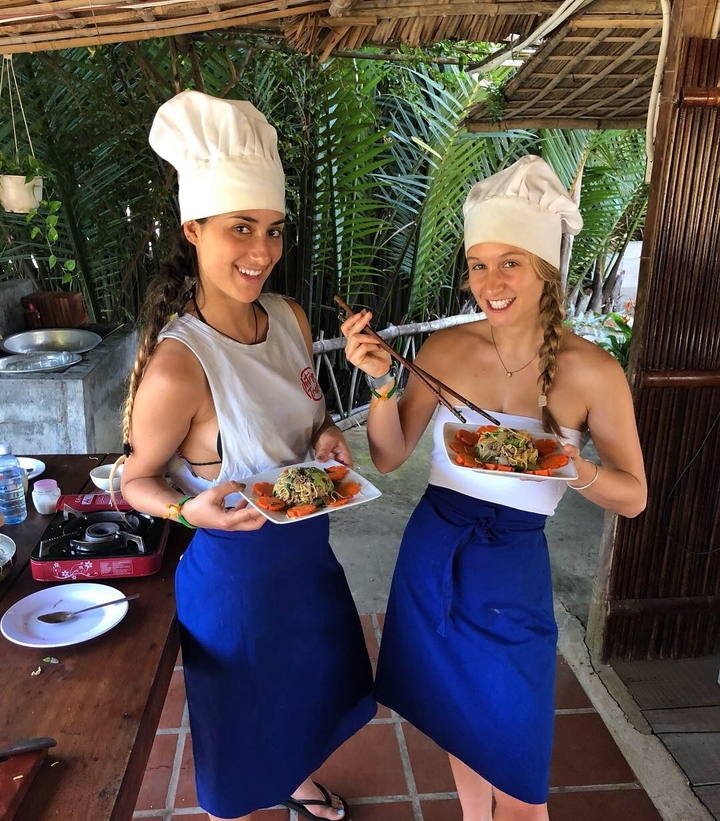 Two women presenting plates of food outside, smiling while holding chopsticks.