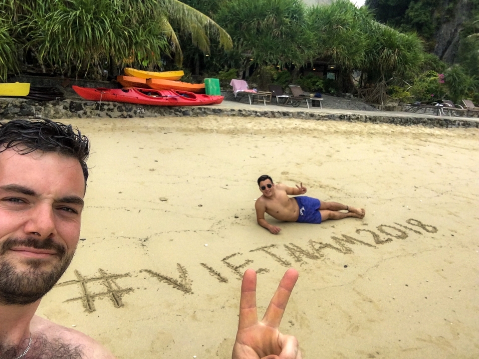Person laying on a beach with writing in the sand, '#Vietnam2018'.