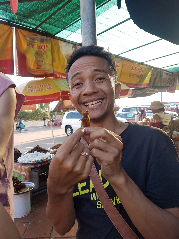Man holding a cooked insect with a happy expression at a market.