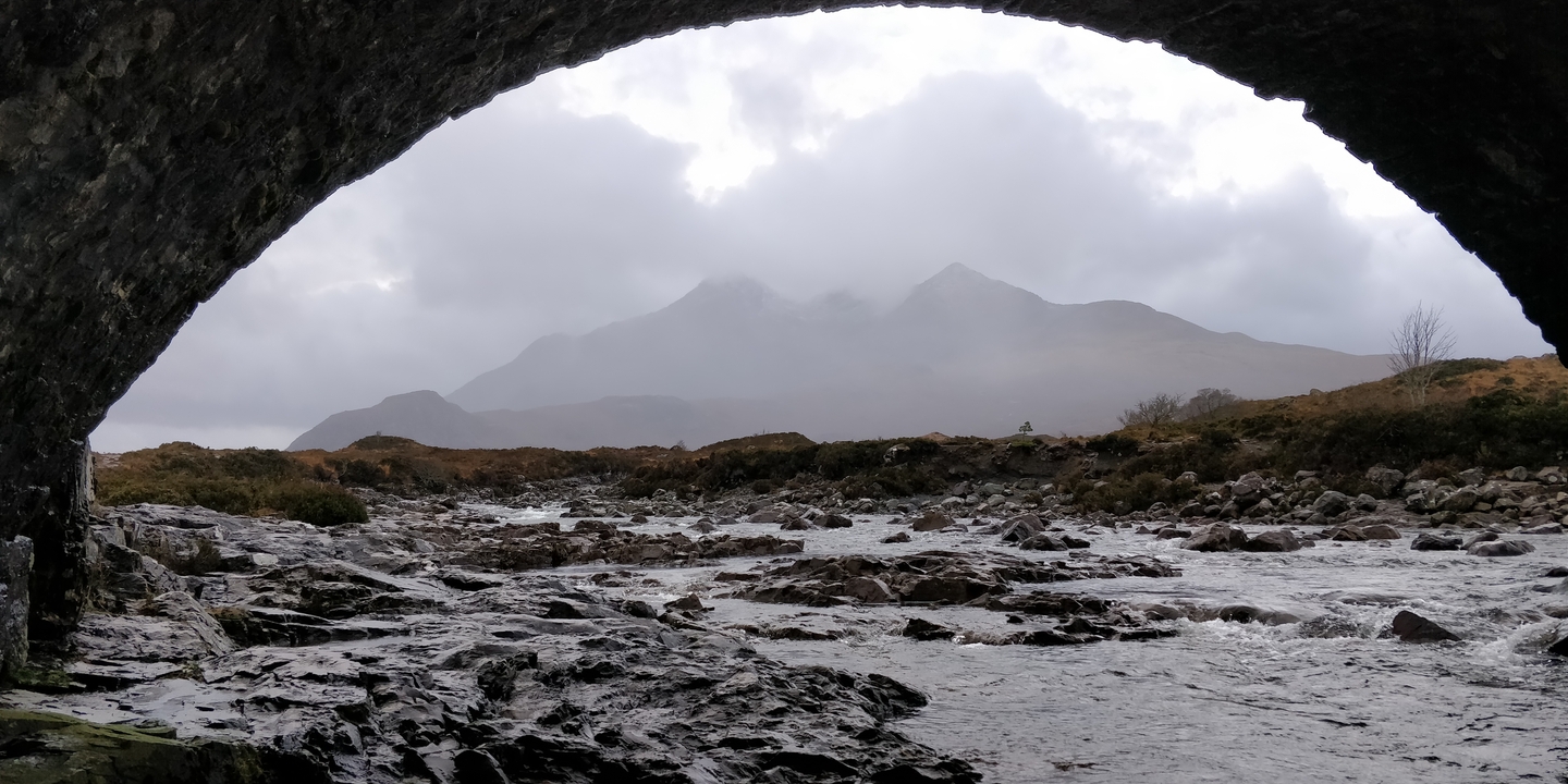 A rocky river flows under a stone bridge with mountains in the background.