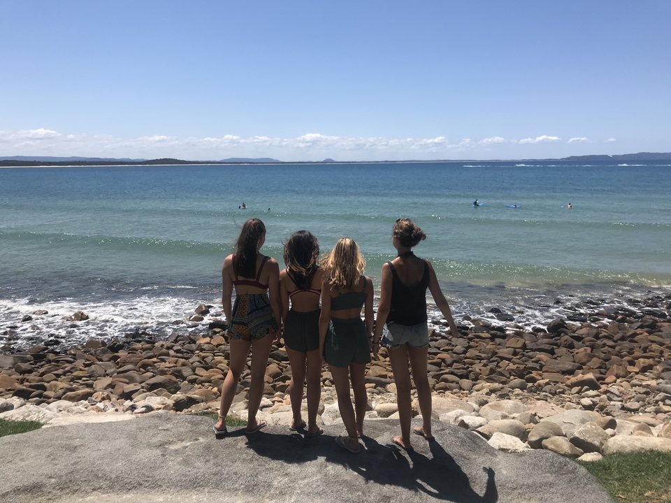 Four women standing on a rocky beach facing the ocean