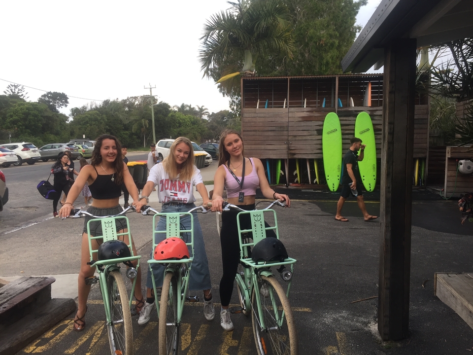 Three women standing with bicycles near surfboards