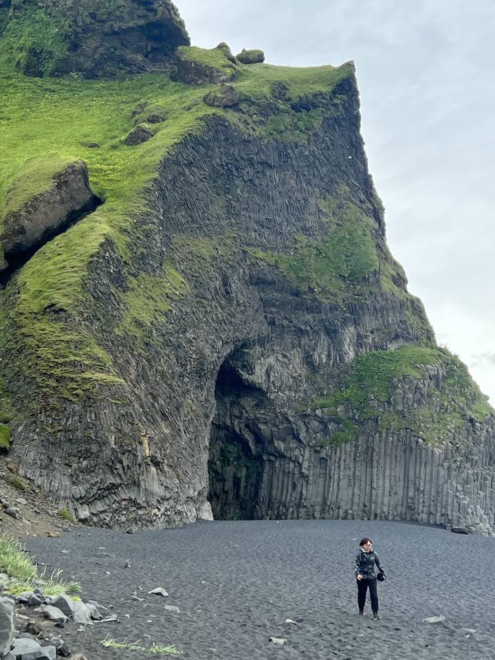 A person standing on a black sand beach with a large moss-covered rock formation.