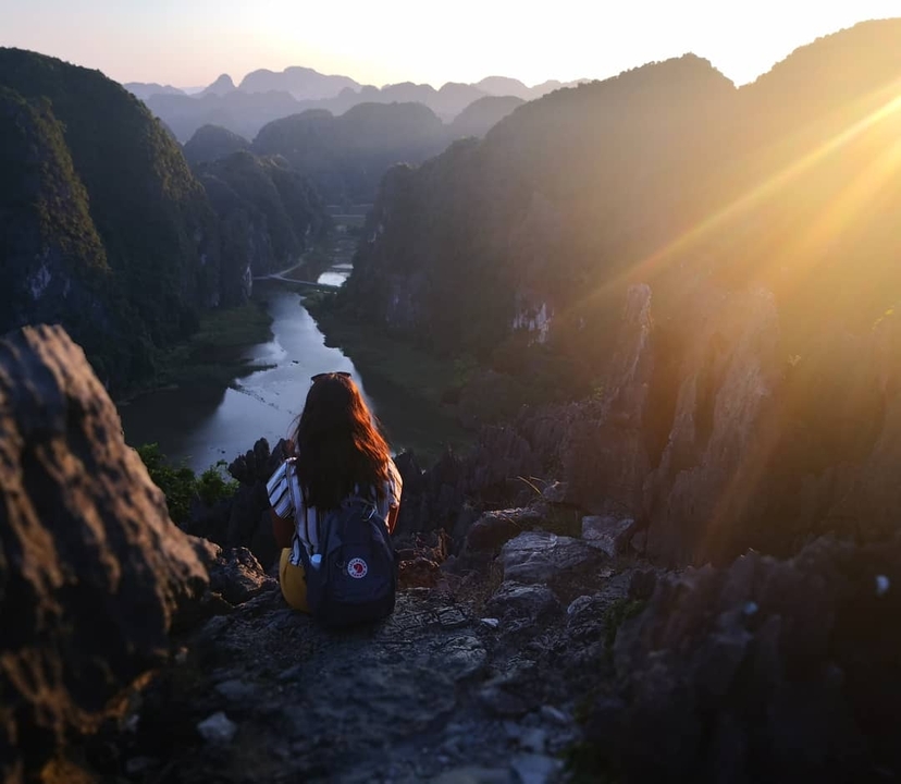 A person sitting on a cliff edge overlooking a river and mountains at sunset.