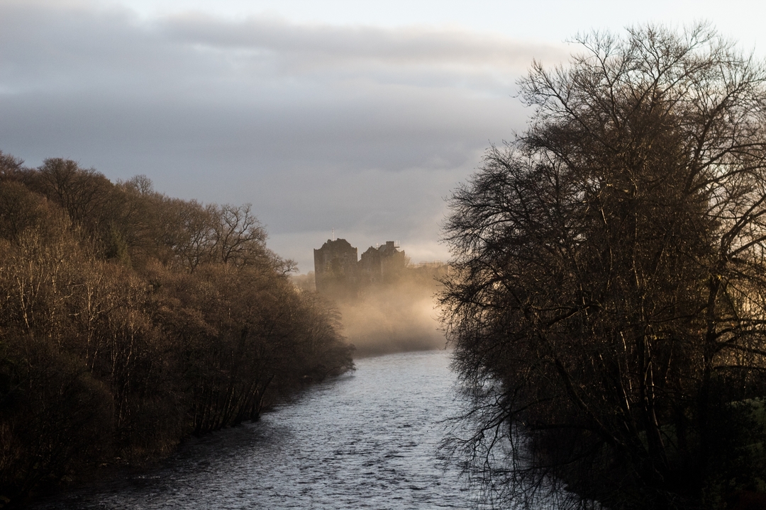 Rivière avec des ruines de château brumeuses en arrière-plan entourées d'arbres.